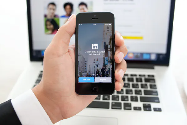 Hand holding an iPhone with the LinkedIn app open in front of a laptop